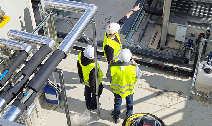 Tre operai con giubbotto ed elmetto di sicurezza ispezionano tubi e attrezzature industriali in un cantiere all'aperto.