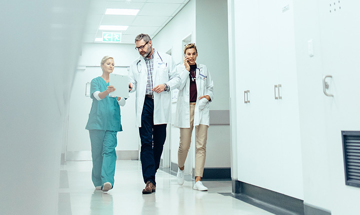 Tre medici professionisti camminano lungo il corridoio di un ospedale, discutendo le informazioni sui pazienti su una tavoletta digitale.