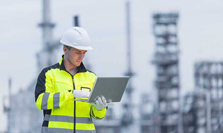 Un operaio edile con giacca ad alta visibilità e elmetto utilizza un computer portatile in un sito industriale.