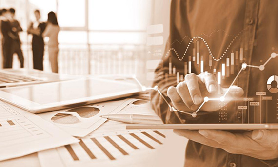 Persona che utilizza un tablet con grafici di dati sovrapposti, documenti e penna su un tavolo, gruppo di persone che parlano sullo sfondo.