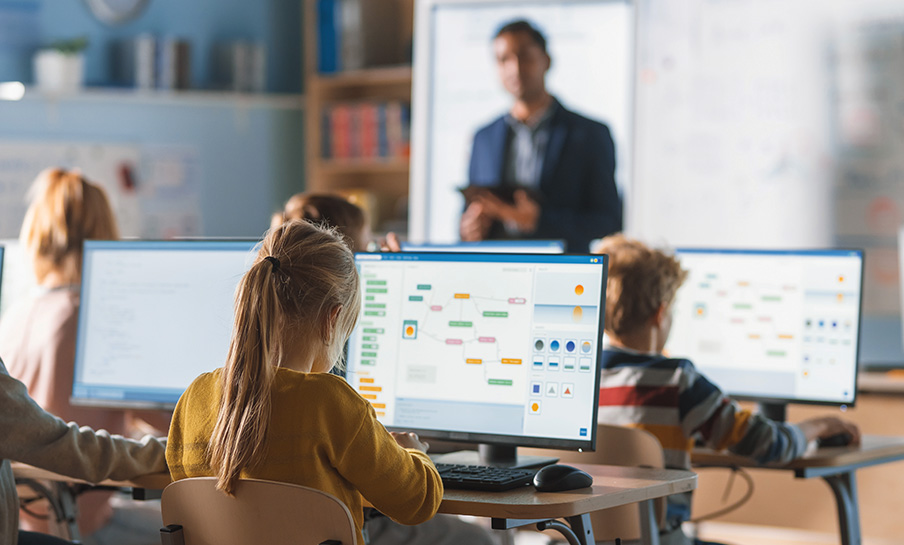 Gli studenti sono seduti ai banchi e utilizzano i computer desktop in un'aula, mentre l'insegnante è in piedi davanti a una lavagna.