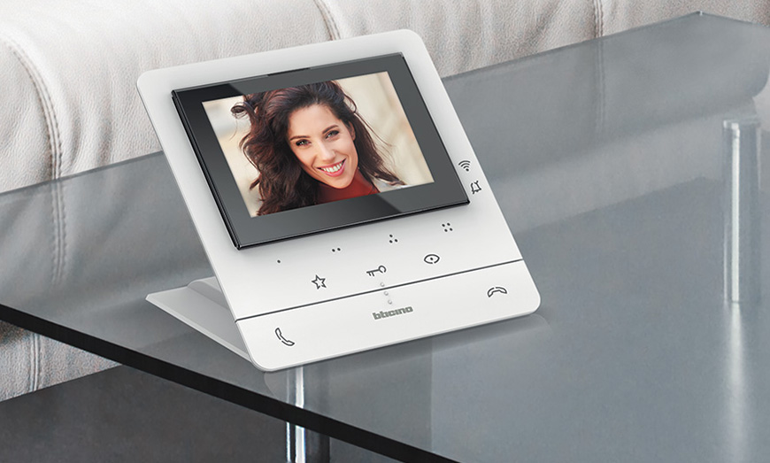 Un videocitofono BTicino Classe 100 con supporto da tavolo è appoggiato su un tavolo di vetro e mostra una donna sorridente sullo schermo.