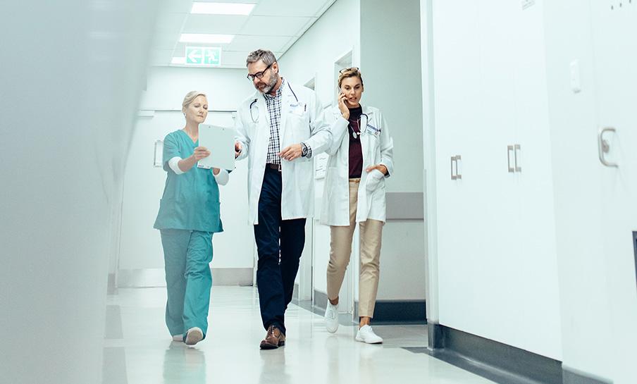 Tre medici professionisti camminano lungo il corridoio di un ospedale, discutendo le informazioni sui pazienti su una tavoletta digitale.